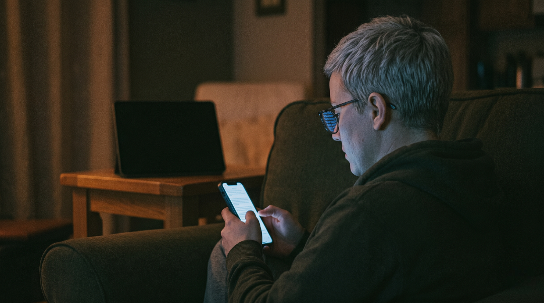 A person seen from behind, sitting alone in a dim room at night, their face faintly lit by a phone screen, with a second screen turned away in the shadows behind them